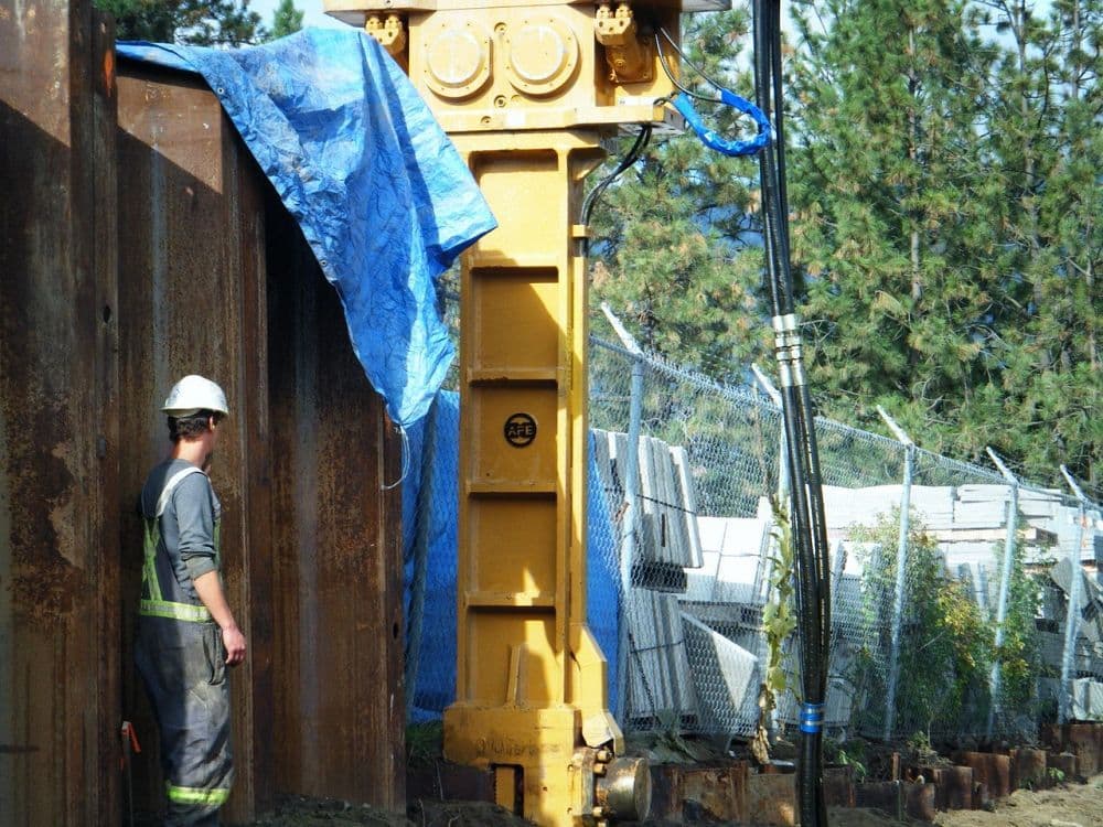 Construction crew guiding steel sheet piling into place at the Craftsman Collision site in West Kelowna, BC, enabling stable foundation work for new building development.