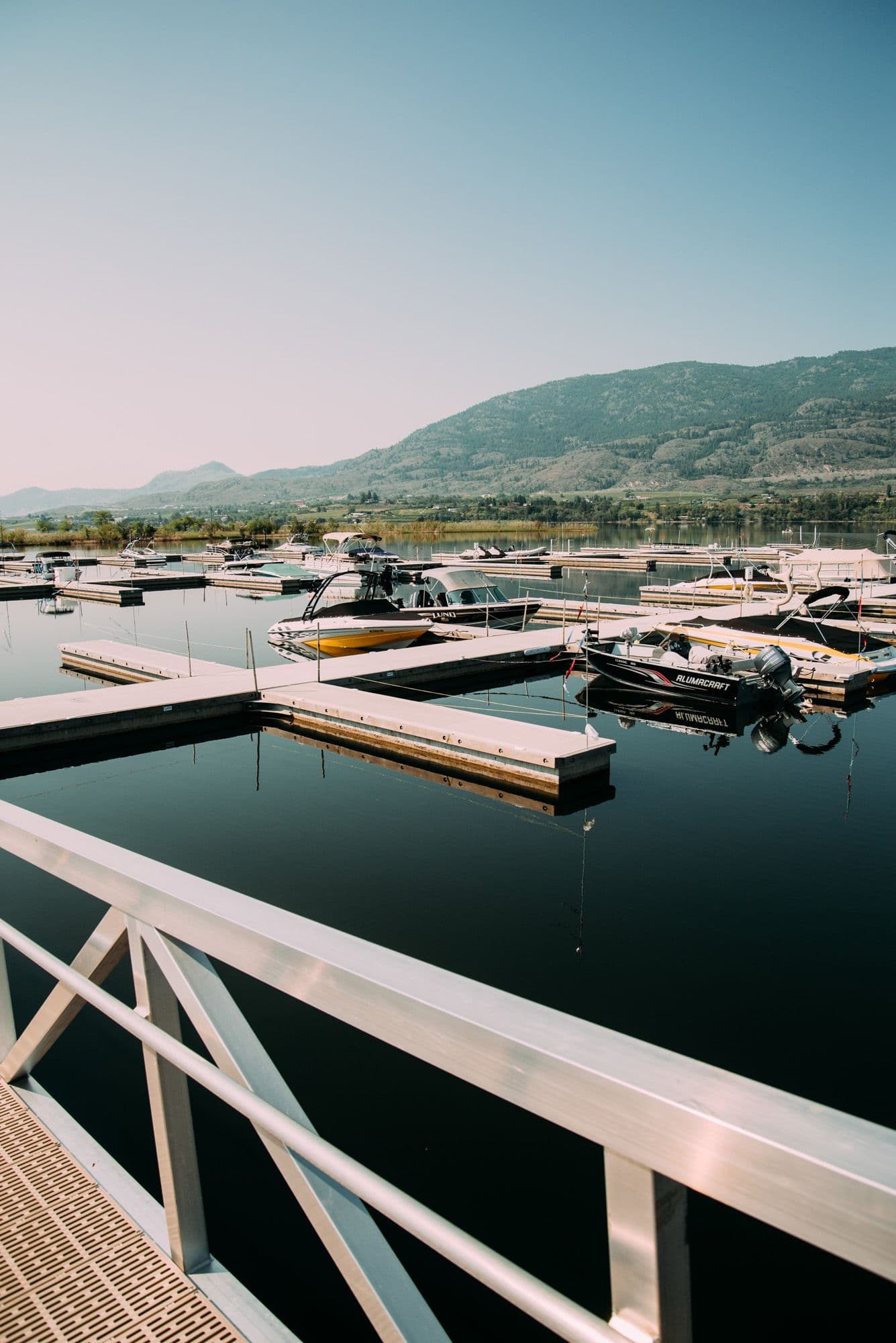 Framed view from marina walkway overlooking boat slips and tranquil water, with Osoyoos hillside in the distance.