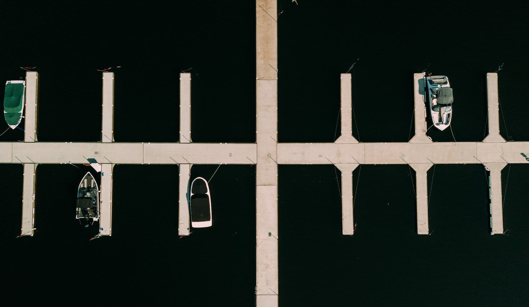 A symmetrical aerial view of a modern boat marina, showing empty and occupied dock slips extending into calm, dark water. Several boats are moored in clean, evenly spaced berths