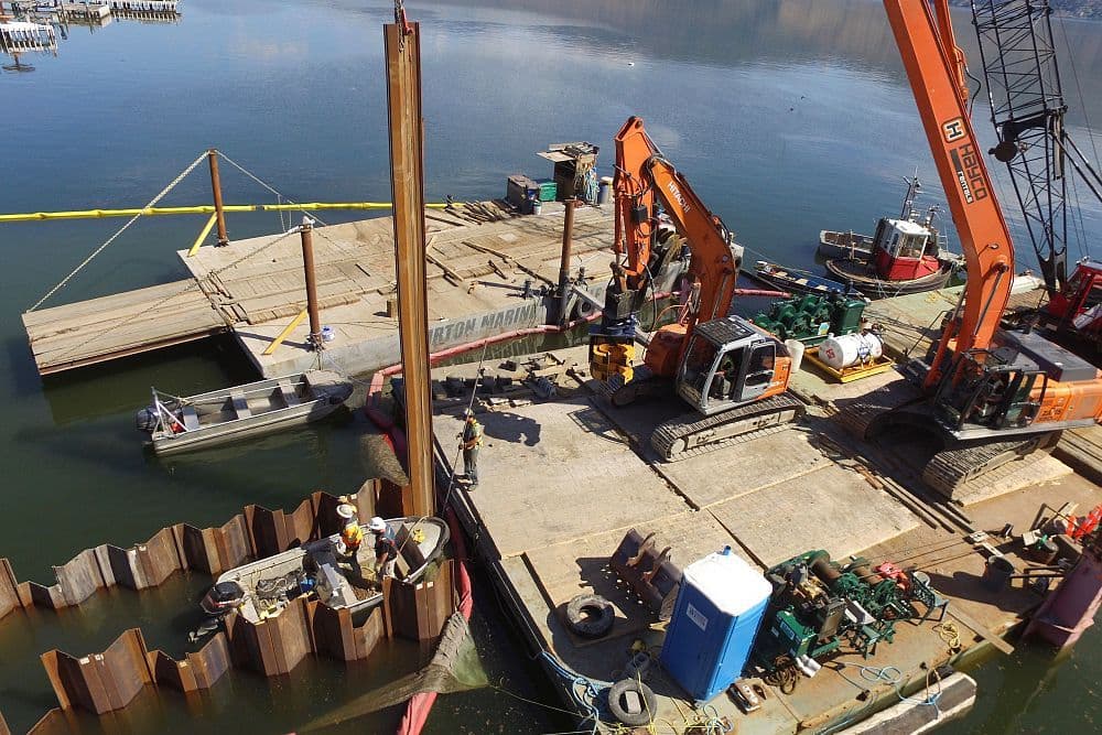 Excavators and barges on Okanagan Lake installing a temporary sheet pile wall for RDCO sewage outfall pipeline protection in West Kelowna, BC.