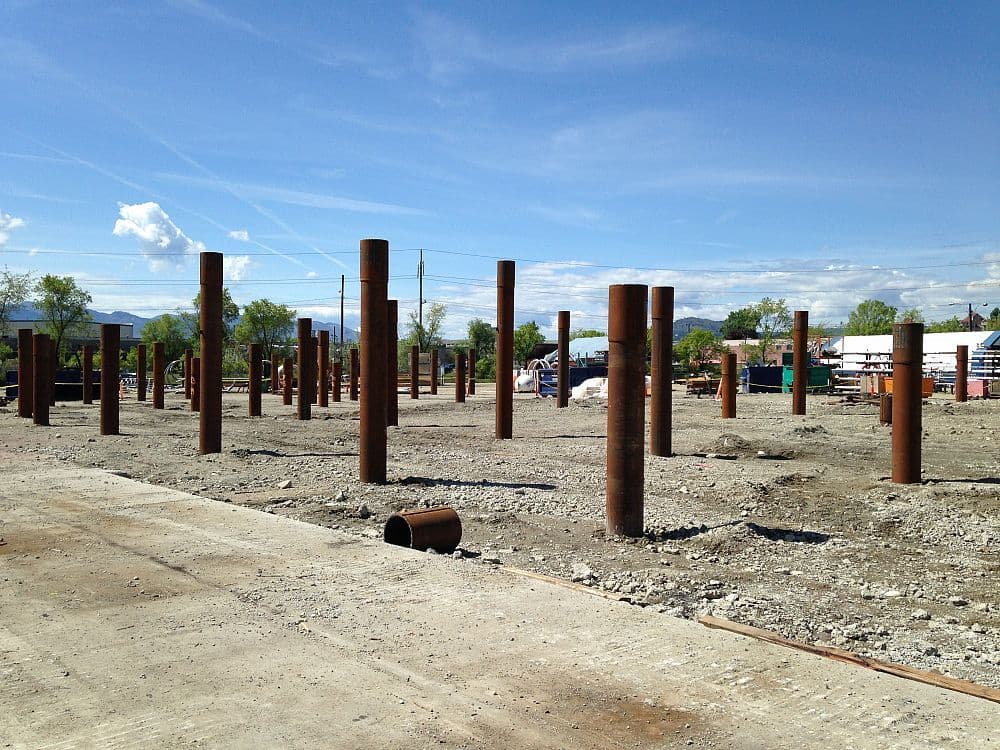 View of a cleared construction site with foundation piles driven into the ground, surrounded by gravel and concrete surfaces.