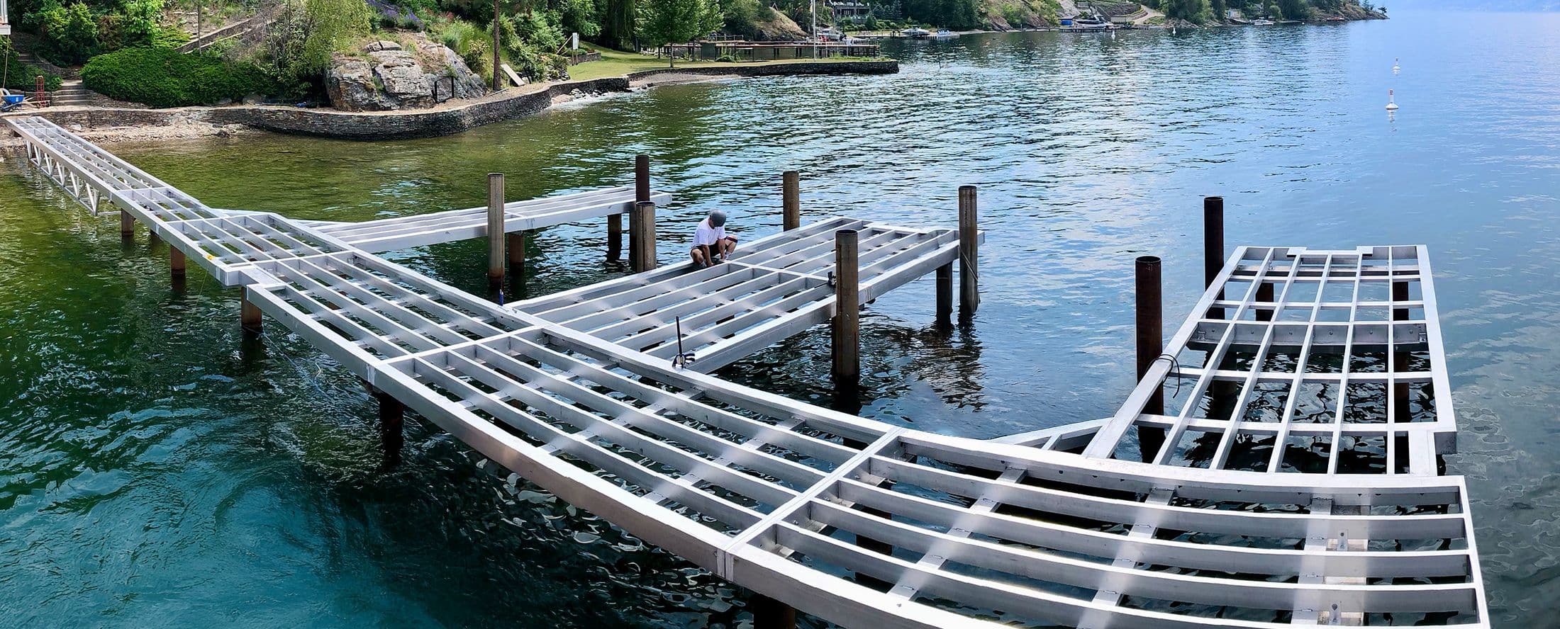 A worker installing a marine-grade aluminum dock frame over water, surrounded by cylindrical pilings in a clear lake setting.