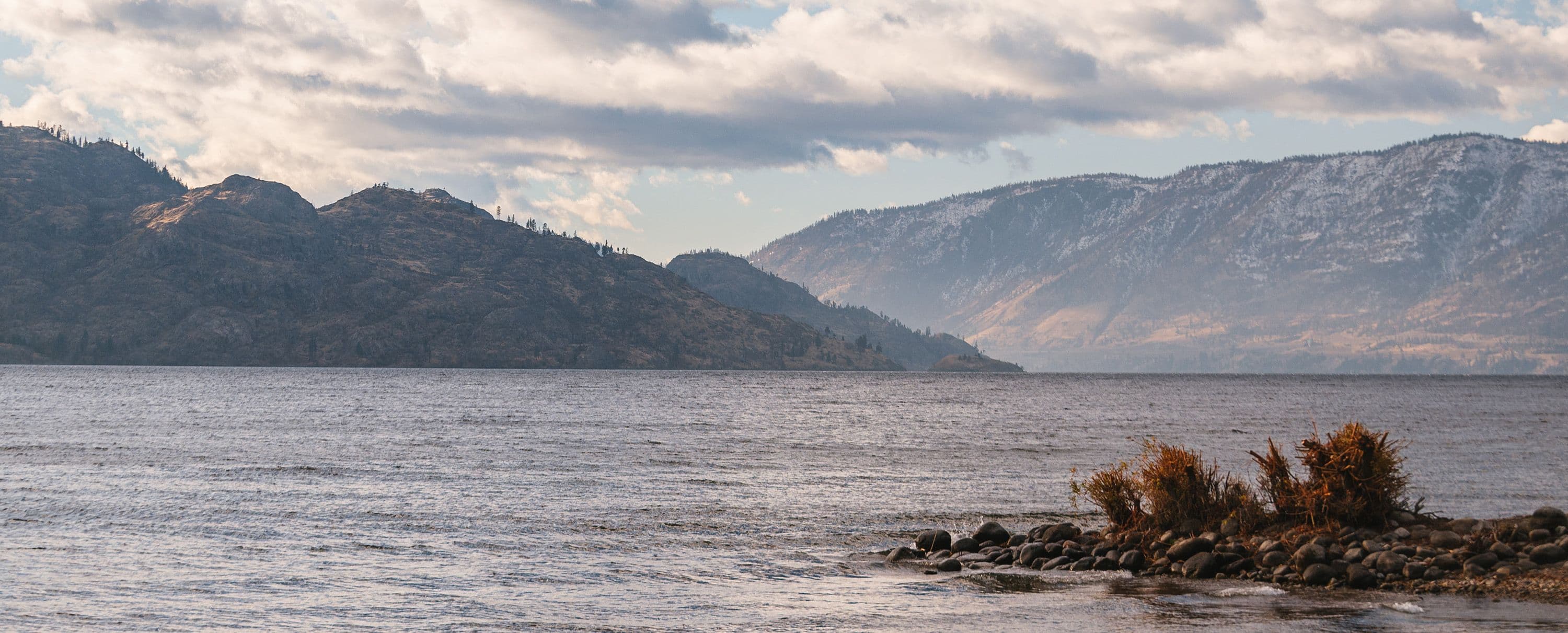 Wide view of Lake Okanagan under a partly cloudy sky, with mountain ranges in the background and rippling lake water in the foreground. The landscape conveys natural beauty and a sense of expansive tranquility.