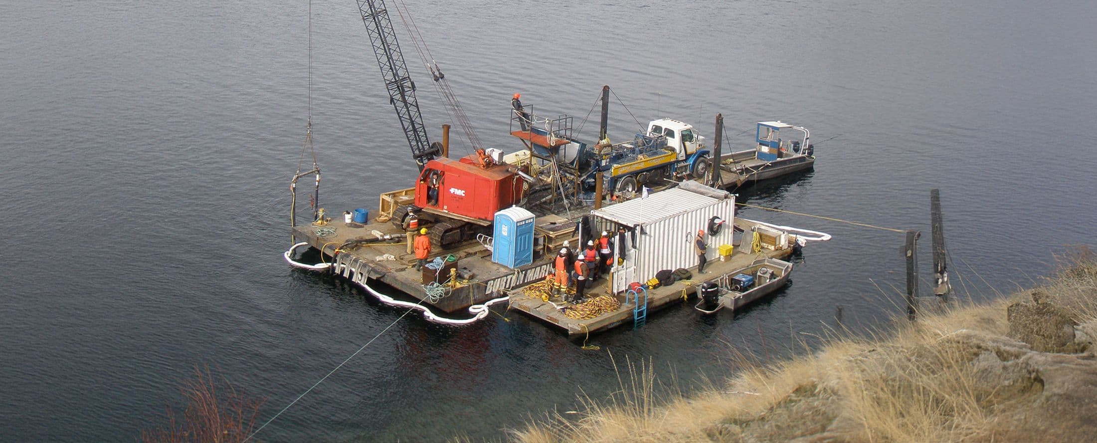 Floating barge loaded with marine service equipment and crew, including a crane, truck, and work containers, performing waterfront operations on a calm body of water.