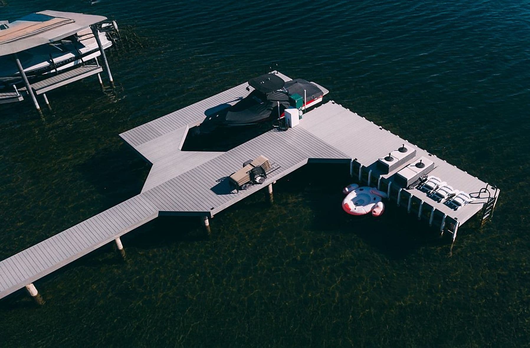 A modern, light-colored dock extending over clear water, featuring a bench, a boat with a canopy, and a round inflatable float tied to the side.