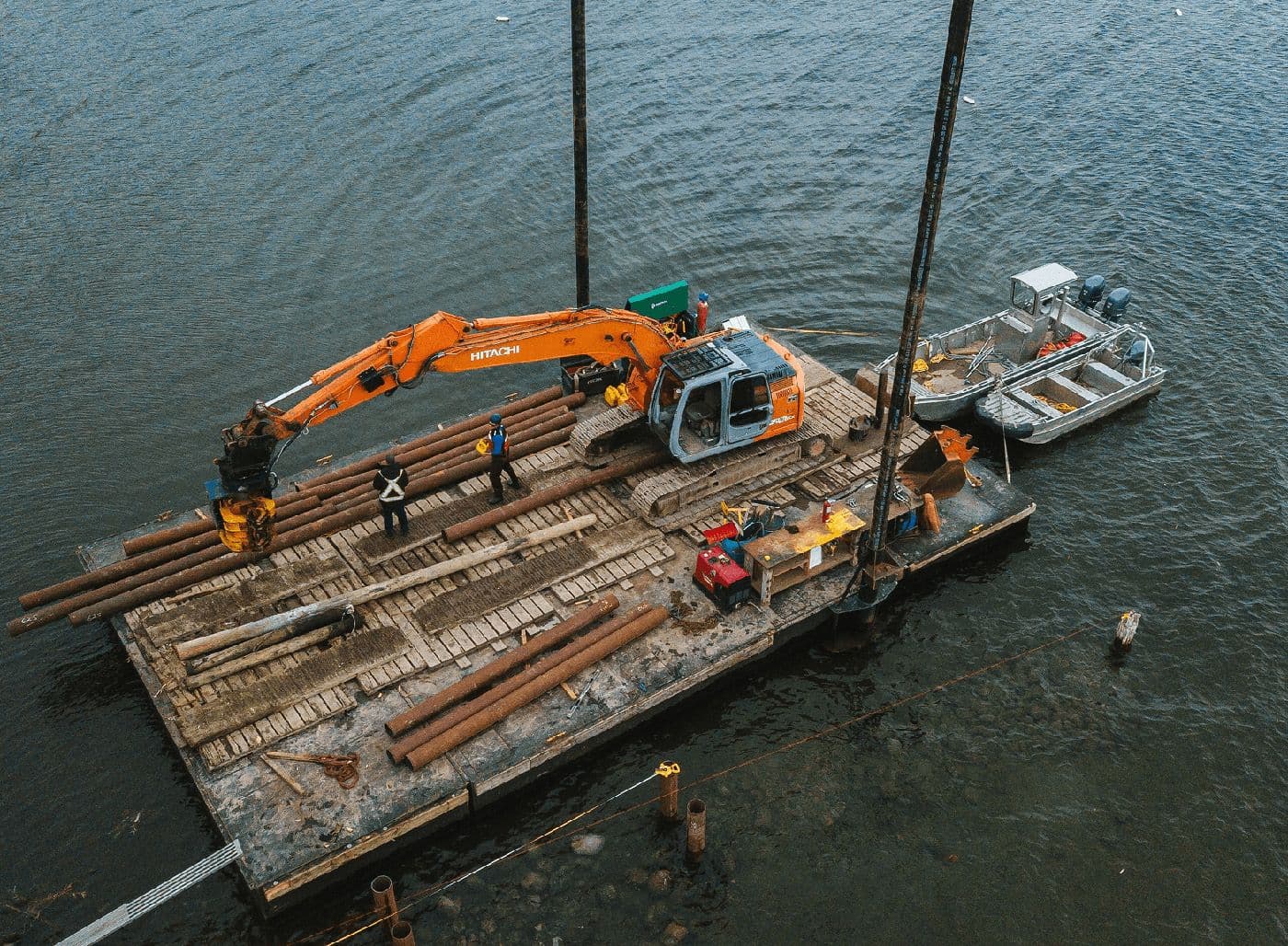 A construction barge with an orange excavator and two workers dismantling and loading large wooden beams from an old dock on a body of water.