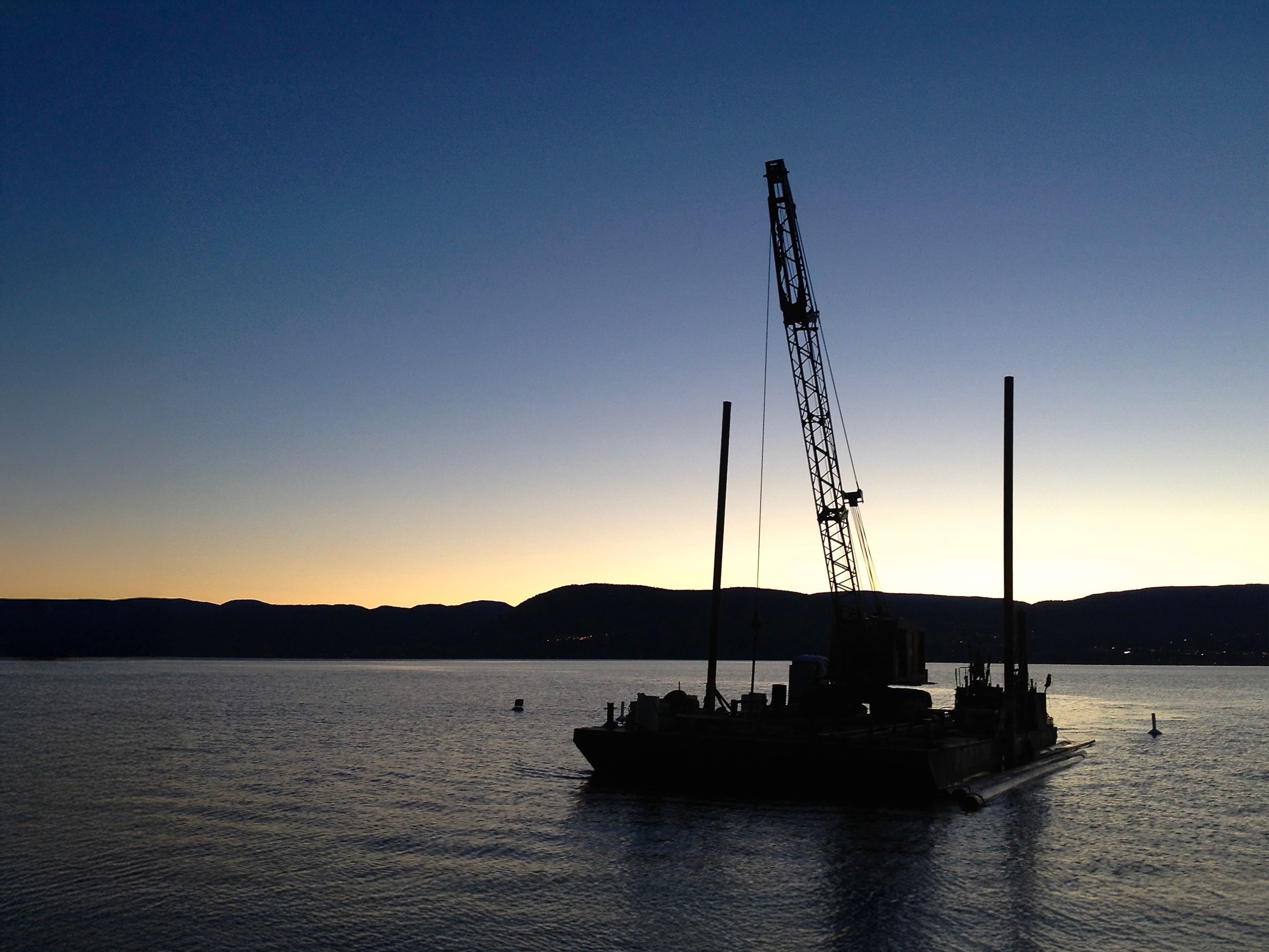 A crane barge silhouetted against a lake at dusk, with soft twilight over the mountains and a calm water surface reflecting the fading light.